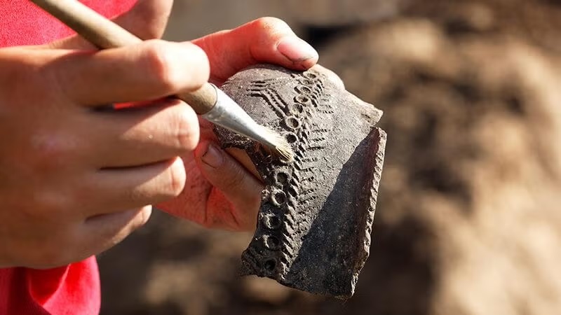 hands holding a tool and an artifact over dirt.