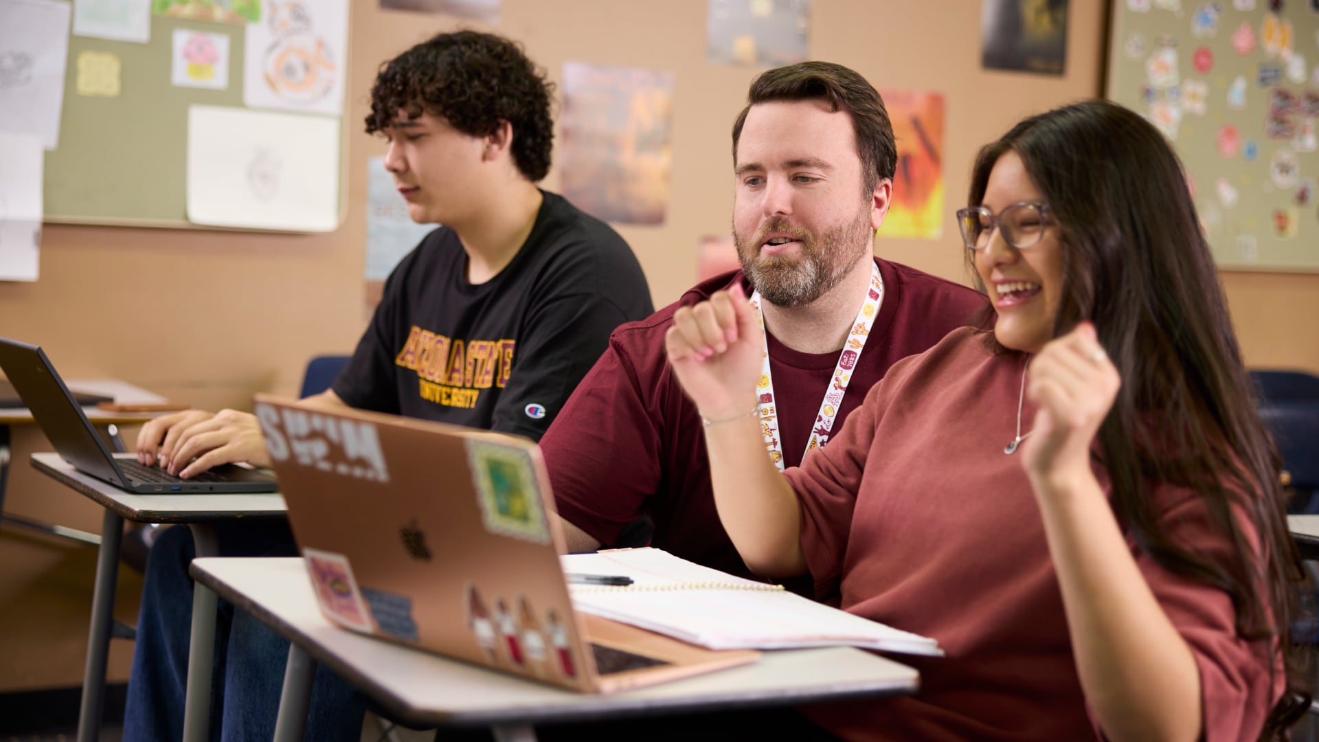 A teacher and two high school students work on laptops in a classroom, smiling as they collaborate on an English course assignment.