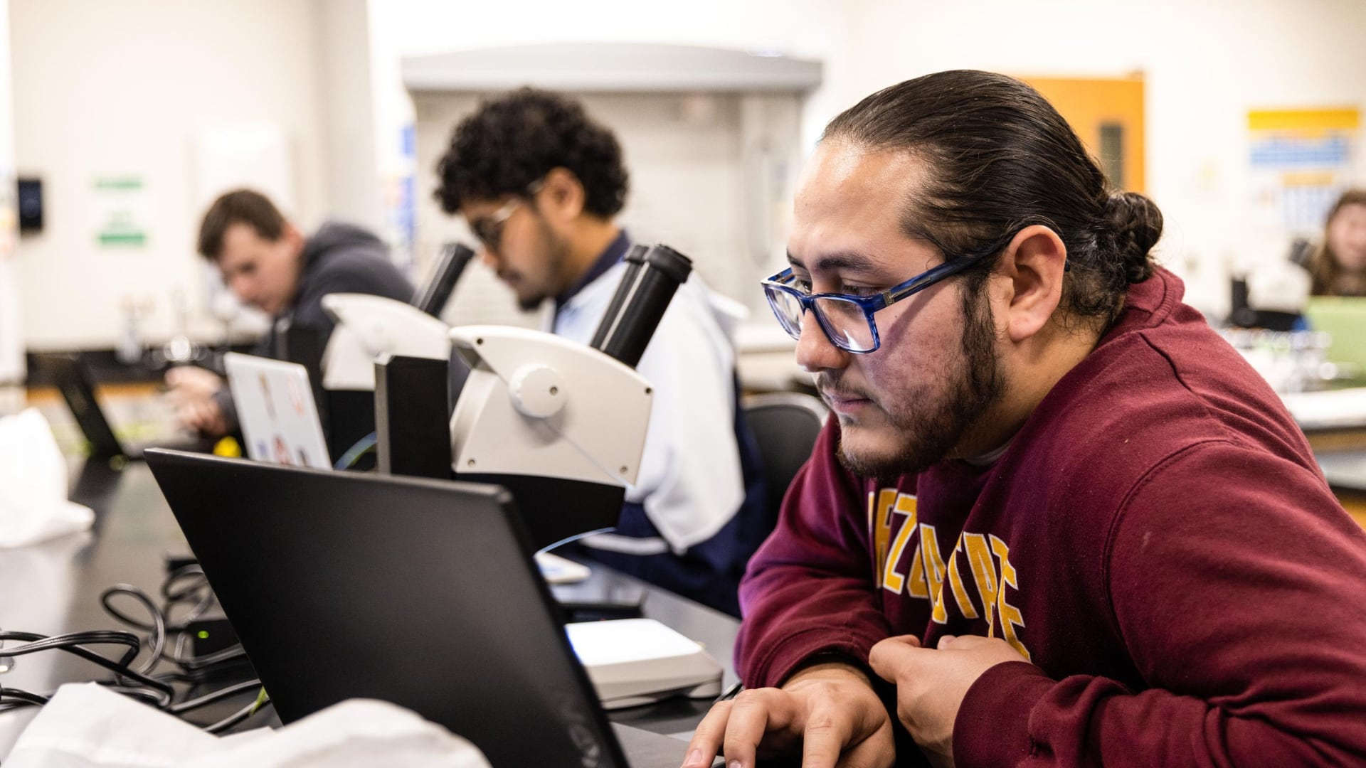A student in a science lab works on a laptop beside a microscope, using curated educational resources while classmates study in the background.