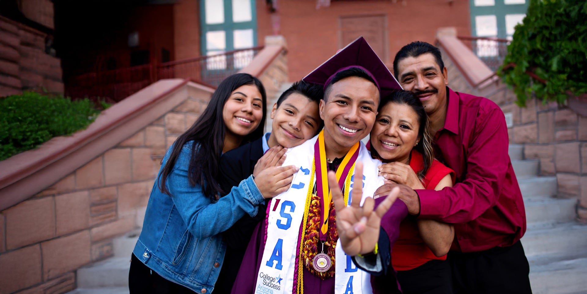 A graduate in cap and gown smiles with family embracing him on the steps of a building, celebrating his earned admission and achievement.