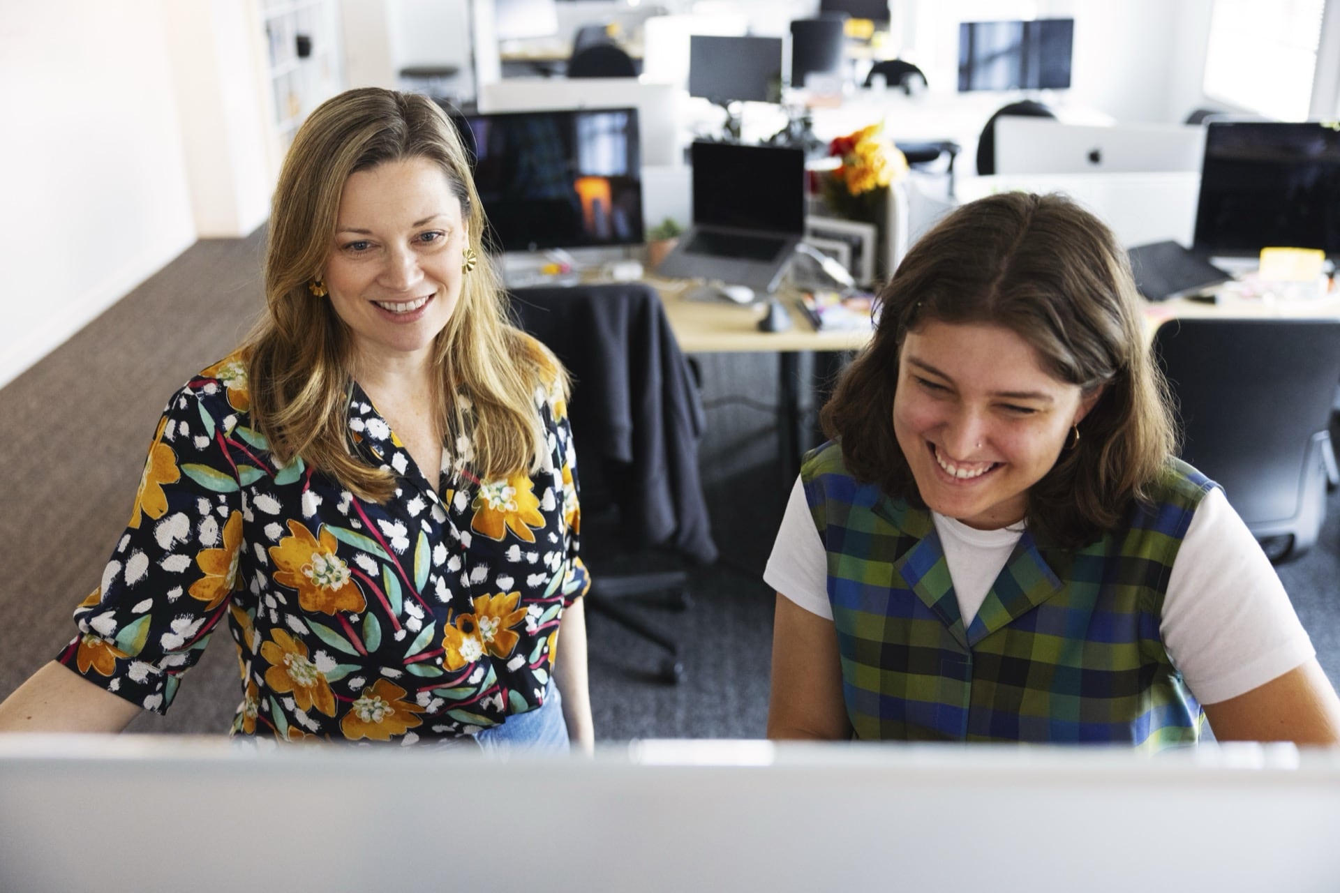 Two colleagues stand together smiling at a computer screen in a bright office, reflecting supportive leadership and collaborative management.