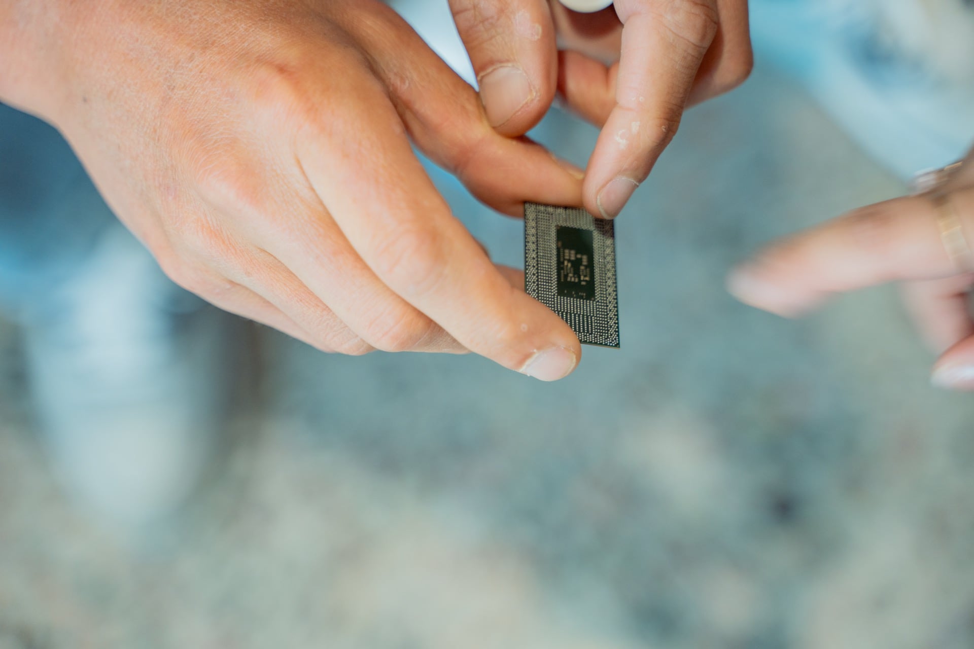 Hands holding a small microelectronics chip while another hand points toward it against a blurred background.