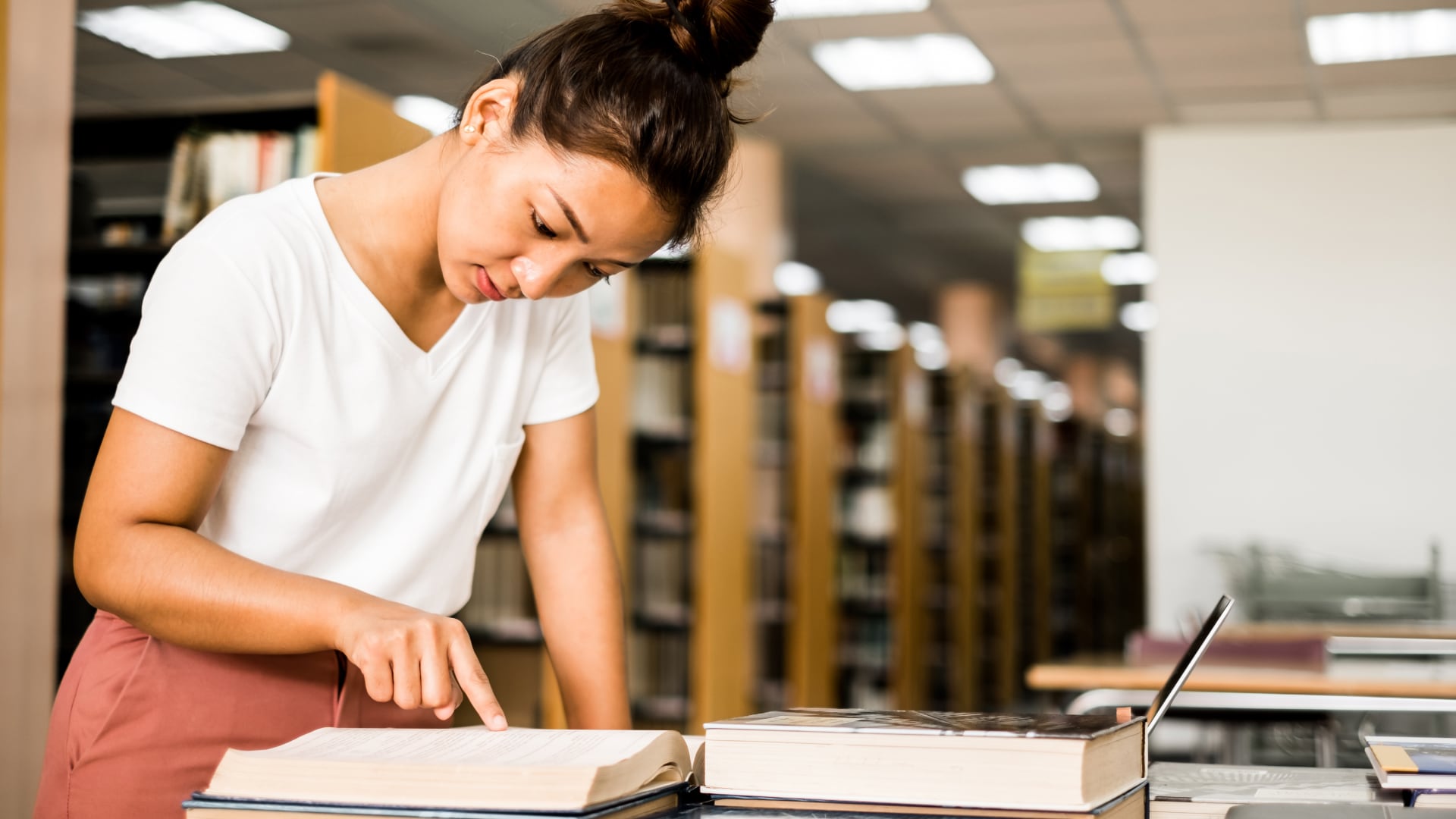 A student in a library leans over an open book, pointing to a passage while studying modern history.