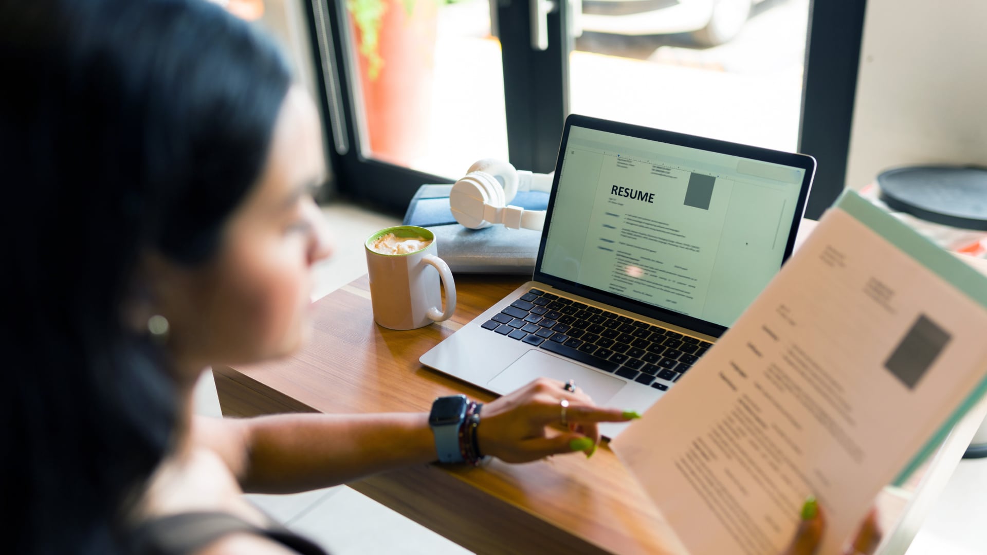 A woman reviews a printed resume while comparing it to a digital version on her laptop in a café workspace.