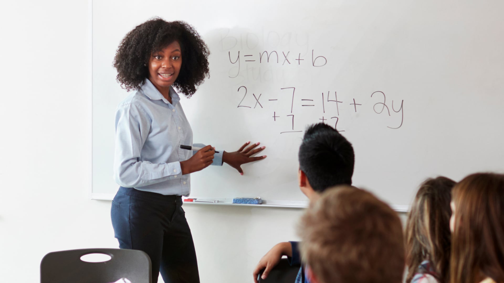 Teacher writing algebra equations on a whiteboard while students watch, illustrating a classroom lesson in algebra and precalculus.