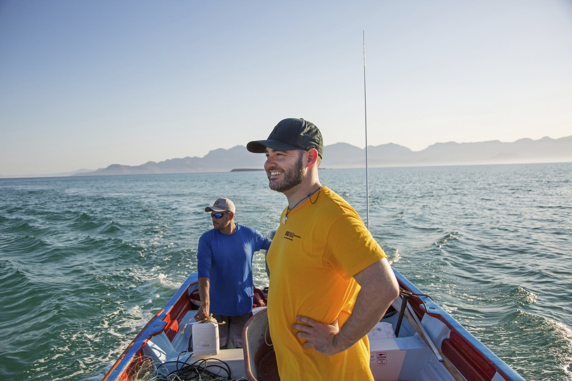 Two people stand on a small boat in open water with mountains in the distance, highlighting fieldwork connected to sustainability.
