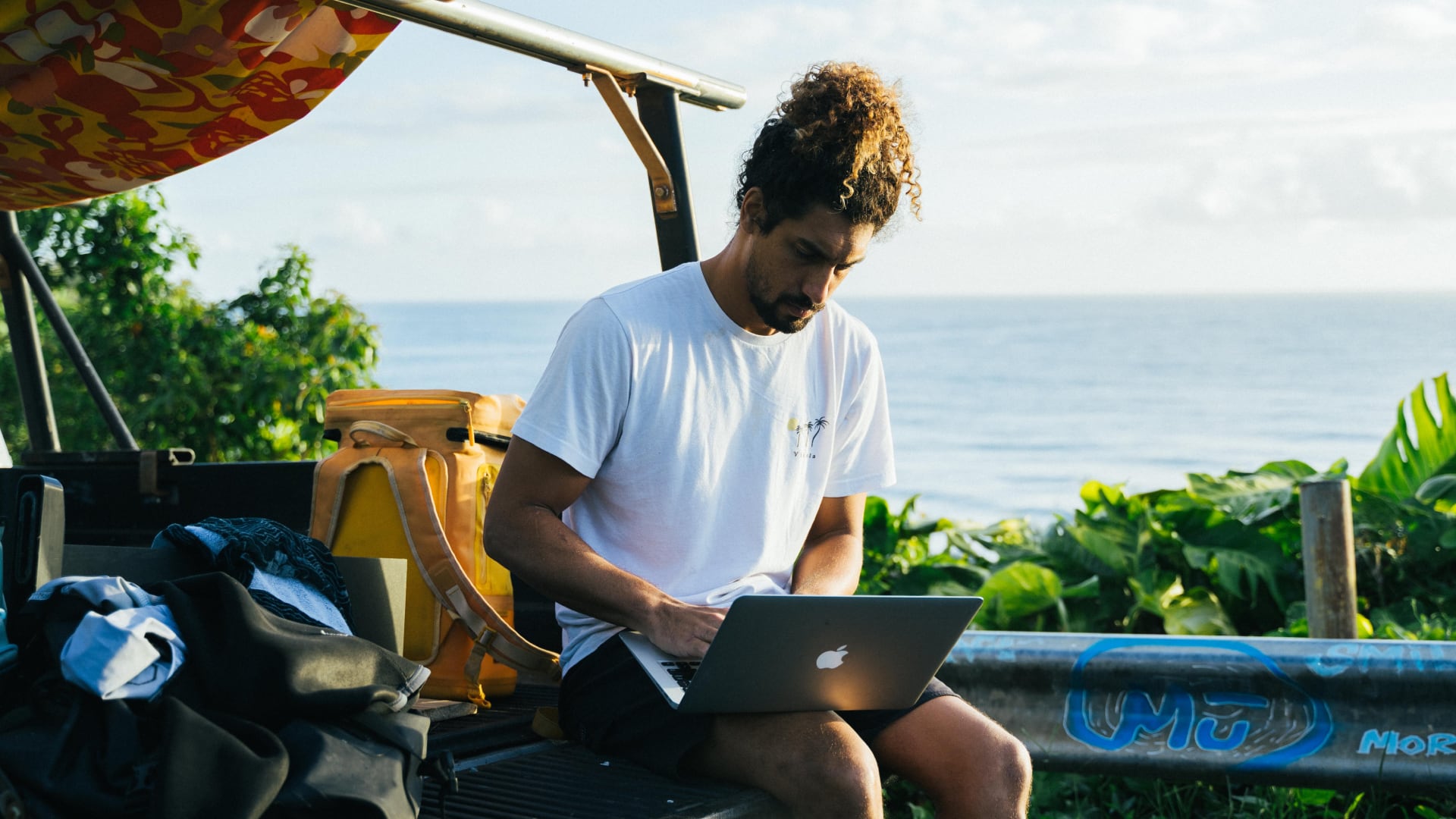 A man works on a laptop beside the ocean with teaching materials nearby, illustrating sustainability lessons for teachers in an outdoor setting.