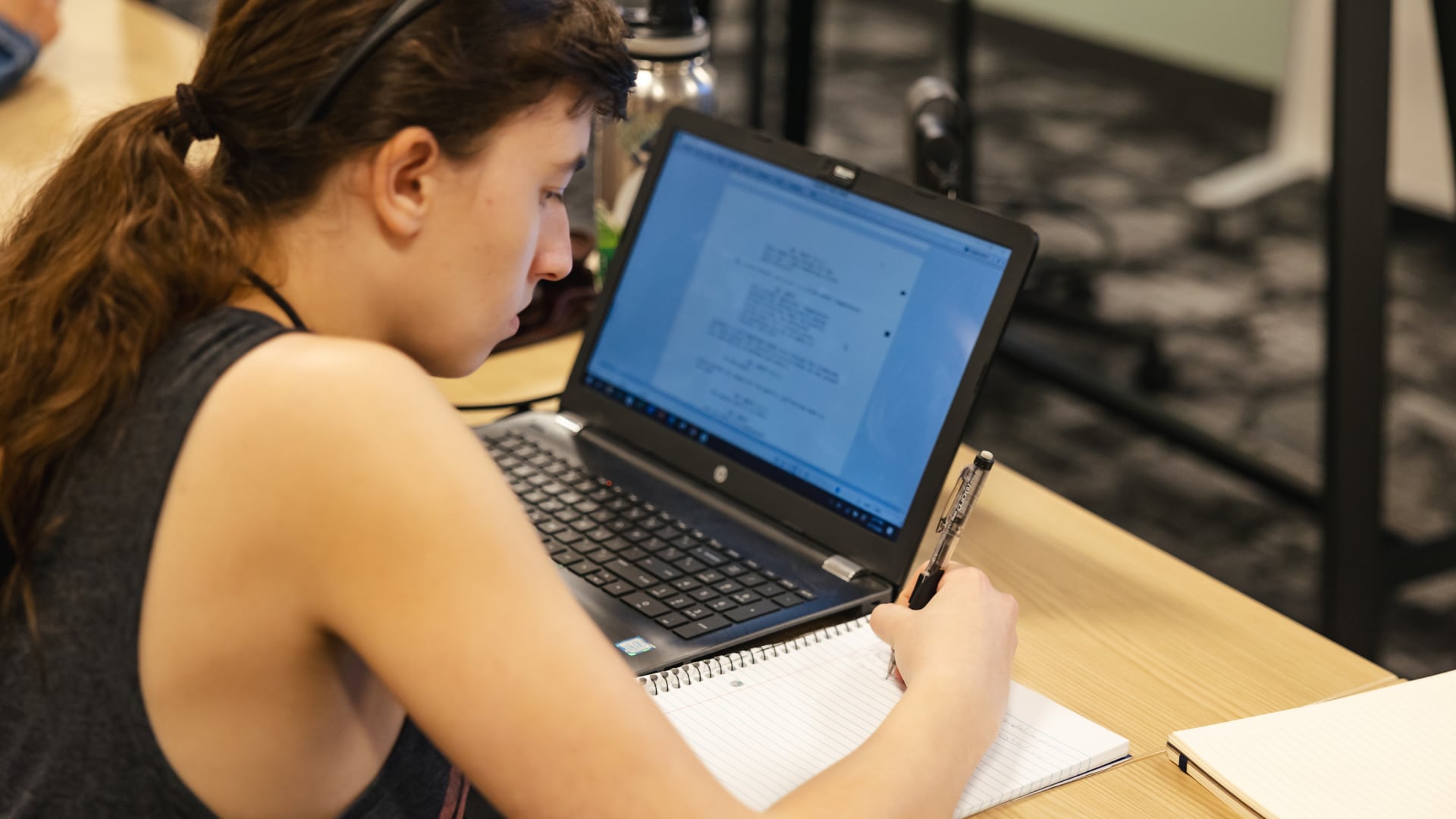 A student writes notes in a notebook while reviewing text on a laptop during a writing composition class.